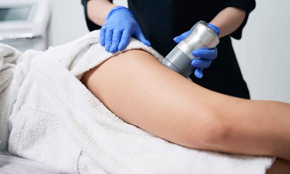 Aesthetician performing a cellulite treatment on a woman's thigh using a handheld device in a clinic setting.